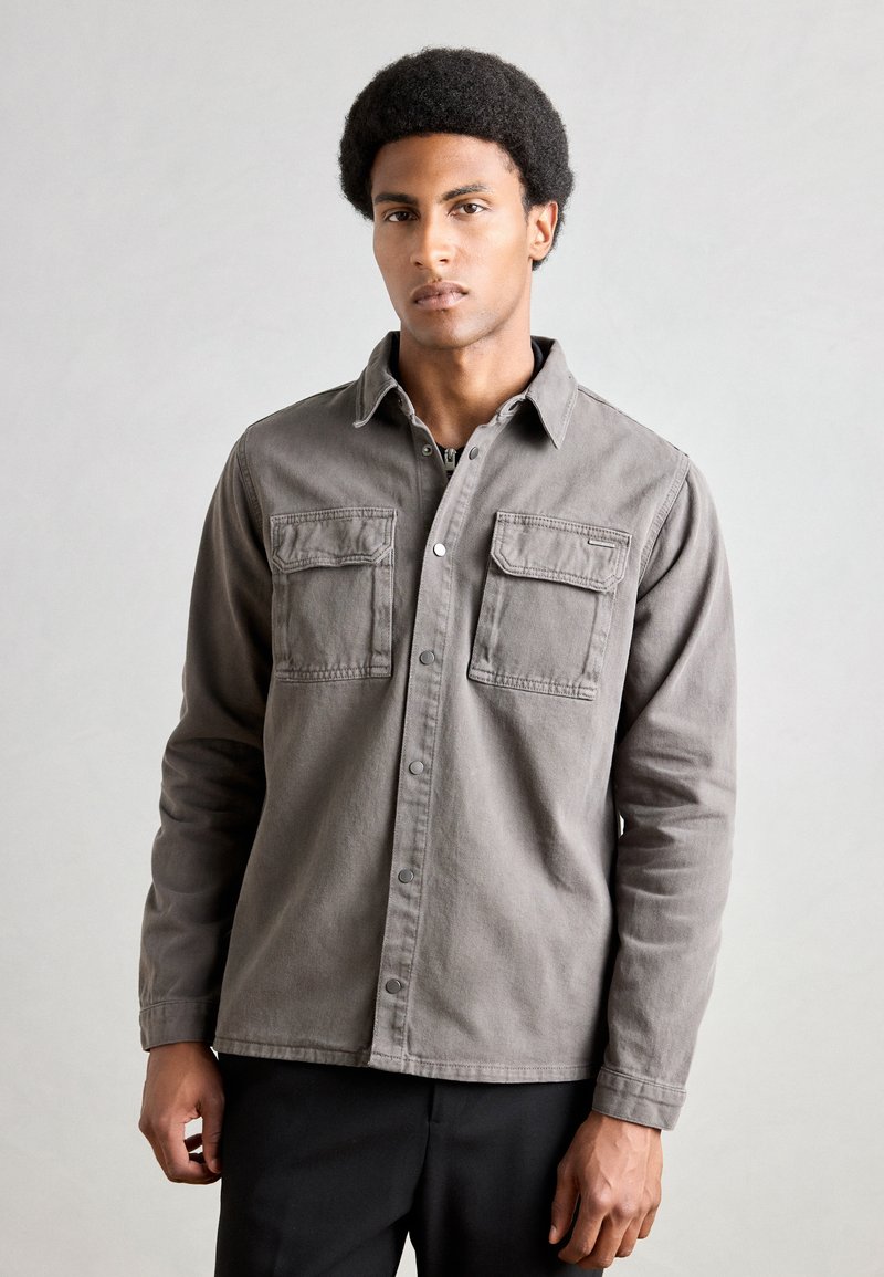 Camisa gris de botón, hecha de una tela resistente, con dos bolsillos en el pecho, un cuello y un dobladillo recto. Corte ajustado con mangas largas.