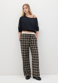 Pull noir à épaules dénudées, design court. Pantalons à carreaux amples en noir et beige avec une taille élastique. Bottines noires à semelle épaisse.