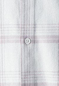 White plaid shirt with light purple grid pattern, featuring a white button and soft, textured fabric. Visible stitching detail along edges.