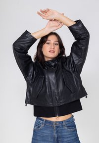 Schwarze lederne cropped Jacke mit hohem Kragen, großen Fronttaschen und Reißverschlussdetails. Getragen über einem schwarzen Oberteil und kombiniert mit blauen Jeans.