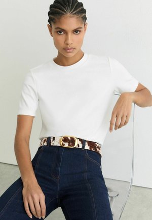 Junge Frau mit geflochtenen Haaren, die ein weißes kurzärmeliges Hemd, dunkle Jeans und einen Gürtel im Kuhfellmuster trägt und auf einem transparenten Stuhl sitzt.