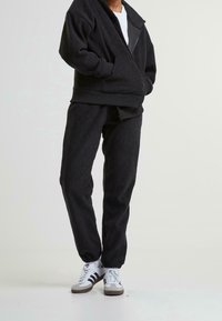 Person in schwarzer Steppjacke mit Reißverschluss und passender Jogginghose, trägt weiße Adidas Sneaker vor weißem Hintergrund.