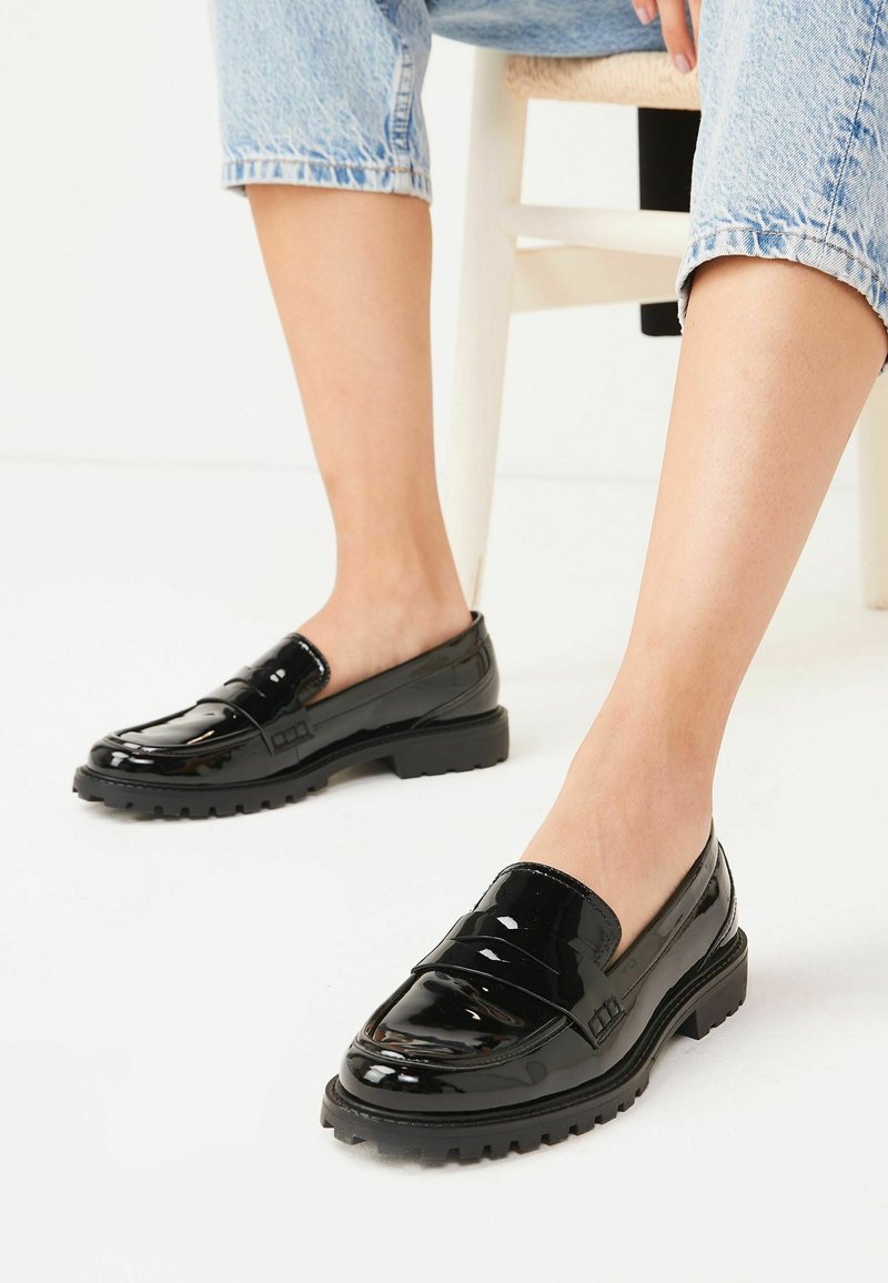 Personne portant des mocassins en cuir verni noir et un jean bleu clair retroussé, assise sur une chaise blanche devant un fond blanc.