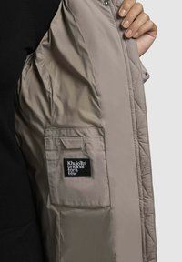 Ruka drží otvorenú béžovú bundu, ktorá ukazuje vnútornú kapsu s čiernou štítkom nesúcim nápis "Khujo TM original for a few" na sivom pozadí.