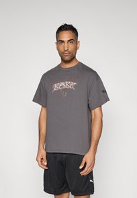 Grå bomulls-T-skjorte med korte ermer, med et grafisk trykk i hvitt og rødt på fronten og en liten logo på ermet. Brukt sammen med svarte shorts.