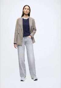 Καφέ και μπεζ σακάκι με μοτίβο καρό houndstooth πάνω από μπλε-σκούρο πουκάμισο, σε συνδυασμό με χαλαρά γκρι τζιν και ανοιχτό μπλε αθλητικά παπούτσια. Απλή διακριτική κοσμηματική λεπτομέρεια.