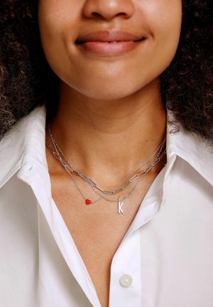 Des colliers en argent superposés avec un charme en forme de cœur rouge et un petit pendentif "k", reposant sur une chemise blanche à col.