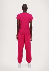 Henkilö seisoo selkä kameraan, yllään kirkas pinkki lyhythihainen paita, siihen sopivat pinkit housut ja valkoiset tennarit yksinkertaista taustaa vasten.