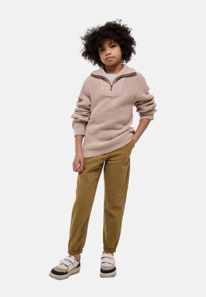 Enfant aux cheveux bouclés debout, une main dans la poche, portant un pull en laine beige, un pantalon marron, et des baskets blanches à scratch.