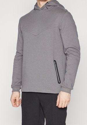 Mann trägt ein graues Kapuzensweatshirt mit einer schwarzen Reißverschlusstasche an der Seite und schwarze Hose vor neutralem Hintergrund.
