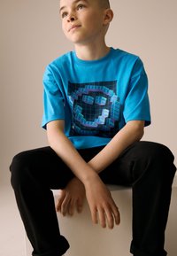 Blauw T-shirt met korte mouwen met een grafisch ontwerp van geometrische vormen op een zwarte achtergrond, gecombineerd met een zwarte broek.