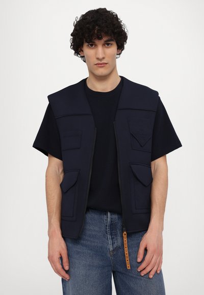 Gilet bleu marine à fermeture éclair, cinq poches, épaules larges et une pochette texturée sur la poitrine. Porté sur une chemise noire et un jean bleu.