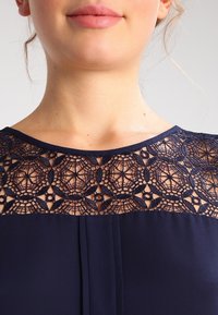 Robe navy avec un empiècement en dentelle décoré de motifs géométriques élaborés, complétée par un tissu lisse en dessous et un col rond.