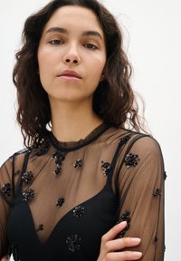 Junge Frau mit dunklem, lockigem Haar, die ein transparentes schwarzes Oberteil mit schwarzen, mit Perlen besetzten, floralem Appliken trägt, die Arme vor einem neutralen Hintergrund verschränkt.