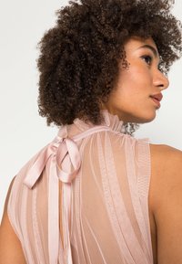 Blusa rosa transparente com textura de riscas verticais, decote alto com babados e laço de fita de cetim nas costas. Aparência de tecido leve e arejado.