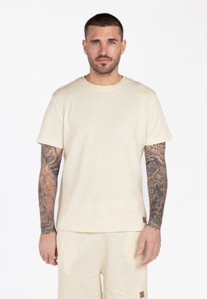 Hombre con tatuajes en ambos brazos, vistiendo una camisa de manga corta texturizada color crema y pantalones a juego, de pie frente a un fondo blanco.