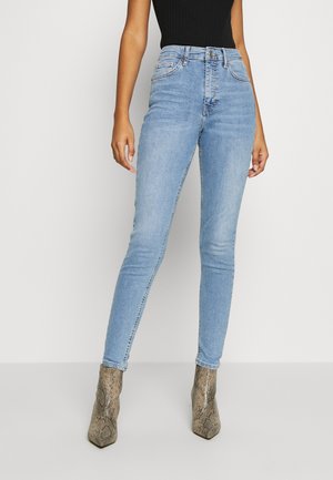 Kvinde iført lyseblå højtaljede skinny jeans, sort ærmeløs top og ankelstøvler med slangeskindsprint og spids snude, stående mod en hvid væg.