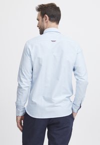 Lyseblå langermet skjorte med hvite vertikale striper, knappe mansjetter og en buet kant. Har et lite merke på baksiden.