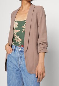 Světle hnědý blazer s volnějším střihem, s klopami a ohrnutými rukávy, v kombinaci s květinovým topem a džíny s vysokým pasem.