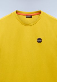 Gul bomulds t-shirt med rund halsudskæring, med en orange detalje ved kraven og et sort og orange logo-patch på fronten.