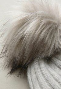 Nærbillede af en hvid strikket hat med en stor, fluffy pom-pom lavet af lysebrun og sortspids faux fur.