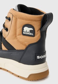 Bota Sorel impermeable con parte superior de nailon marrón, acentos de cuero negro y suela de goma texturizada con una entresuela blanca y detalles de logo.