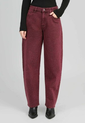 Femme portant un pantalon bordeaux taille haute et ample orné de petites pastilles, un haut noir à manches longues côtelé et des bottes pointues noires.