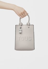 Borsa a tote grigia in pelle con manici arrotondati, logo "TOUS" impresso e un charm rimovibile con tre elementi sferici.
