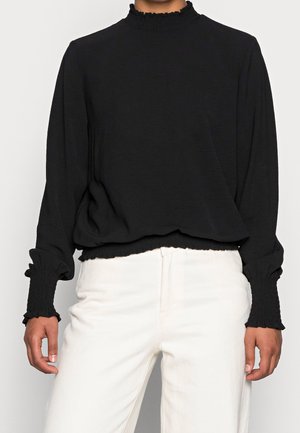 Osoba ve černém topu s dlouhým rukávem s volánkovými manžetami a límečkem v kombinaci s bílými kalhotami, stojící před jednobarevným pozadím.