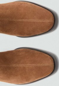 Vue de dessus d'une paire de chaussures en daim beige avec des bouts carrés arrondis et une couture centrale visible sur une surface claire unie.
