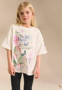 T-shirt surdimensionné beige clair avec des graphiques floraux et animaliers colorés, incluant un oiseau et un papillon, avec une coupe décontractée.