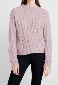 Femme portant un pull en maille épaisse rose clair avec des rayures verticales à motifs, associé à un pantalon noir ajusté.