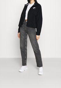 Svart zip-up jacka med Nike Air-logga, vit croppad t-shirt, grå raka jeans och vita sportskor. Slät tyg och avslappnad passform.