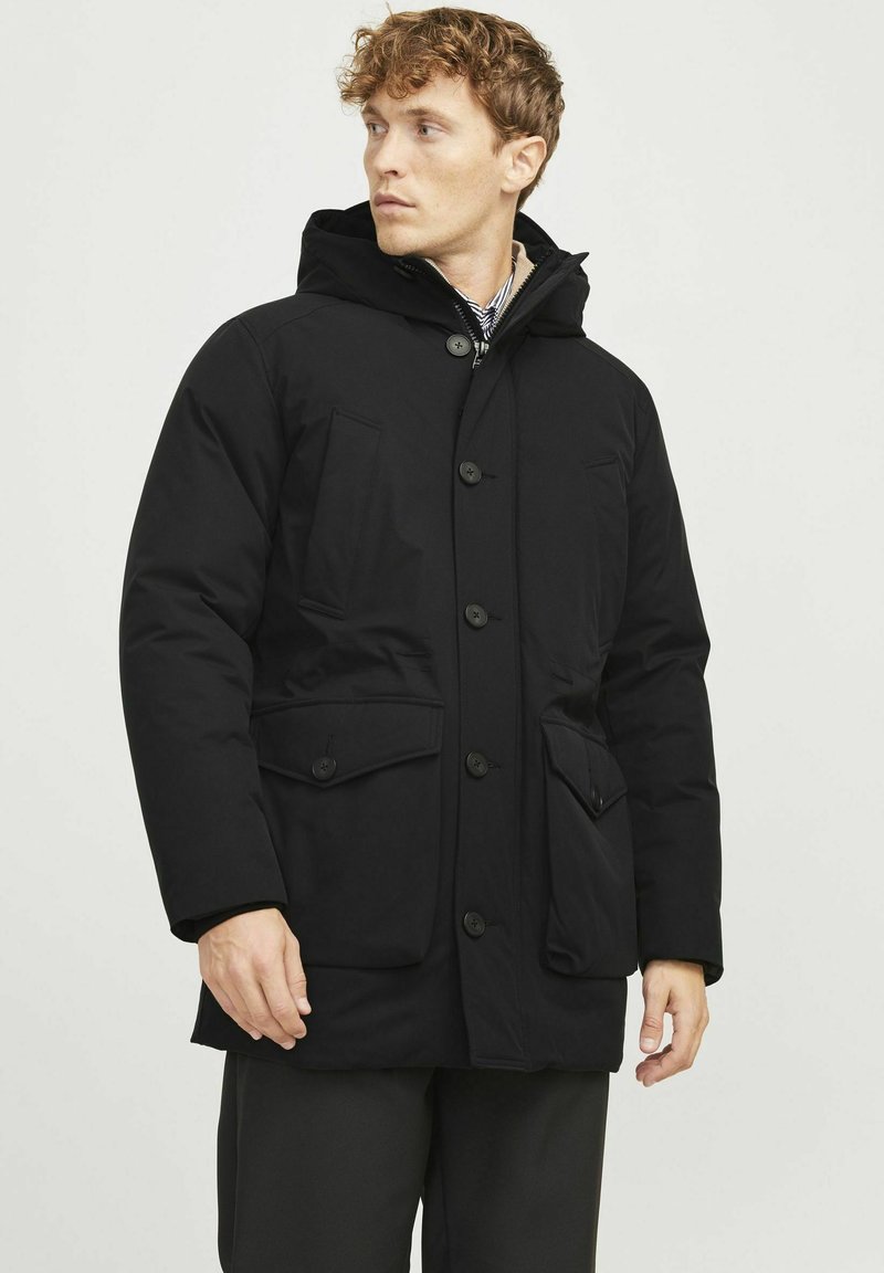 Parka negra aislada con capucha, que cuenta con un cierre de botones, dos grandes bolsillos frontales y puños elásticos, fabricada con un material liso y resistente al agua.
