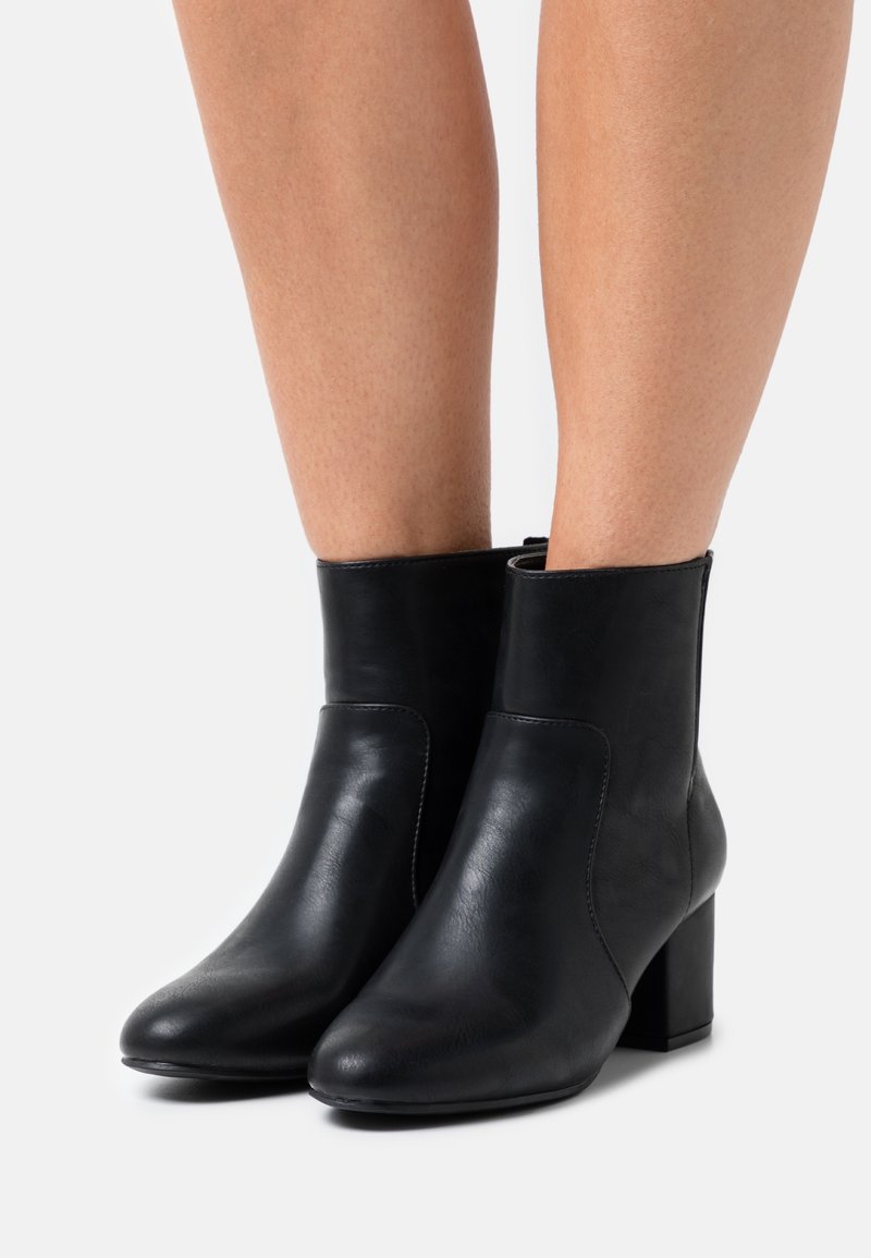 Bottes en cuir noir à talons carrés portées sur des jambes nues, présentées sur un fond uni gris clair.
