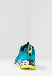Chaussure de sport avec une tige en mesh turquoise, des accents noirs et une semelle blanche et noire à motif. Dispose d'une languette jaune à l'arrière.
