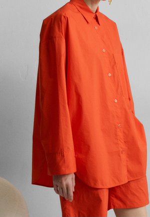Personne porte une chemise orange vif ample à boutons avec manches longues et un short assorti sur un fond gris clair.