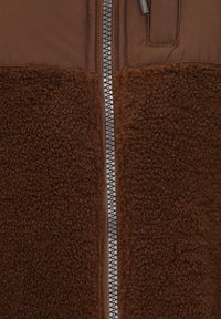 Chaqueta marrón con cremallera, que presenta una sección inferior de forro polar texturizado y suave, y una parte superior de tejido liso, con una cremallera metálica en el centro.