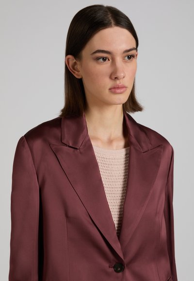 Junge Frau mit glattem braunem Haar, die einen weinroten Satinblazer über einem beigen Stricktop trägt und leicht nach rechts schaut.