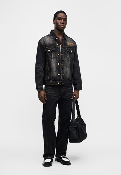 Τζιν μπουφάν και τζιν παντελόνι σε σκούρο πλύσιμο με ξεβαμμένες λεπτομέρειες, χρυσές κεντημένες διακοσμήσεις και μεταλλικά κουμπώματα. Κρατάει μια μαύρη τσάντα. Μαύρα παπούτσια.