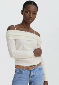 Off-shoulder krem pulover z rebrasto teksturo, dolgimi rokavi in sproščenim krojem, v kombinaciji s svetlo modrim jeansom. Rjav pas z okrasnimi žeblji.