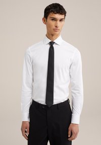 Bijela košulja dugih rukava s crnom kravatom, u kombinaciji s crnim hlačama. Glatka tkanina, klasični ovratnik, prilagođeni kroj, suptilni sjaj kravate.