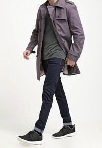 Abrigo largo morado sobre una camisa verde, jeans de mezclilla oscuros y zapatillas negras con suelas blancas. Se pueden ver detalles de botones y charreteras en los hombros.