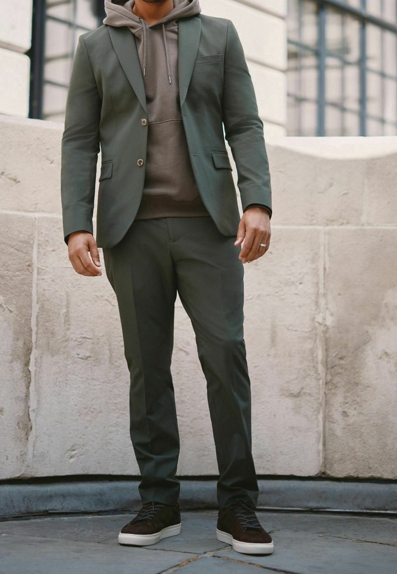 Homme portant un costume vert foncé, un sweat à capuche marron et des baskets noires à semelles blanches, debout à l'extérieur contre un mur en pierre claire.