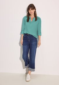 Blusa turchese con scollatura a V, maniche arrotolate e vestibilità ampia, abbinata a jeans alti blu scuro e scarpe da ginnastica bianche.