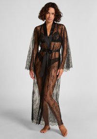 Vrouw staat blootsvoets in een zwarte doorschijnende kanten lange kamerjas met satijnen afwerking, om de taille geknoopt over zwarte lingerie, tegen een effen achtergrond.