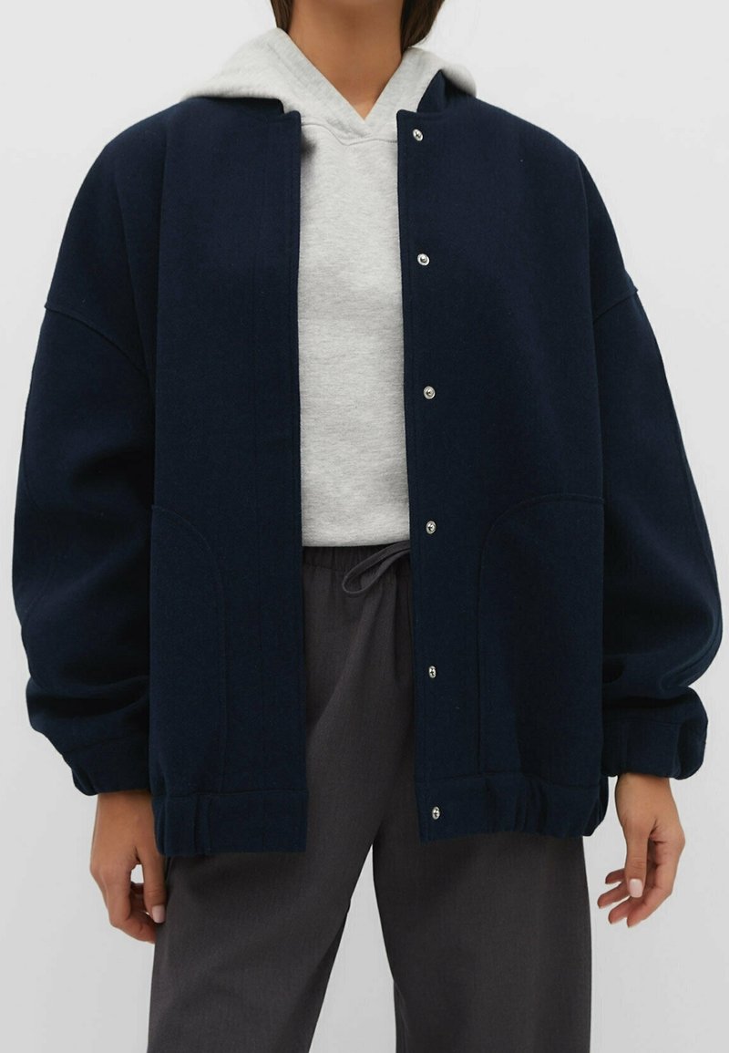 Personne portant une veste bleu marine boutonnée sur un sweat à capuche gris et un pantalon gris foncé à cordon, debout devant un fond uni.