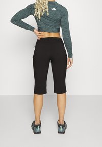 Žena s dlouhými kudrnatými blond vlasy, oblečená v dlouhém rukávovém crop topu, černých 3/4 kalhotách a šedých sportovních botách, stojící zády k nám.