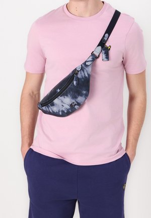 Mężczyzna w różowej koszuli z krótkim rękawem i granatowych spodniach, z niebiesko-białą torbą typu crossbody z nadrukiem tie-dye przewieszoną przez klatkę piersiową i rękami w kieszeniach.
