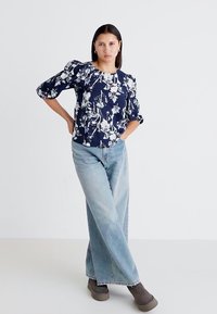 Marinebluse mit Blumendruck und Puffärmeln, kombiniert mit hellblauen, weit geschnittenen Jeans und braunen Knöchelschuhen, vor einem weißen Hintergrund.